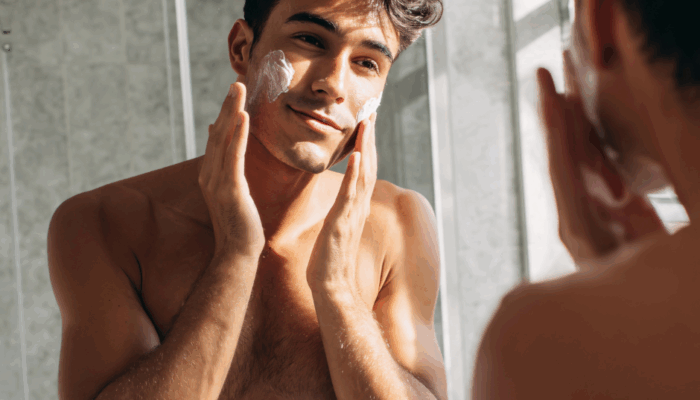 Junger Mann trägt Gesichtscreme auf und betrachtet seine Haut im Spiegel, natürliche Pflegeroutine bei Tageslicht.