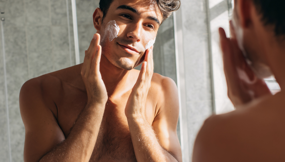 Junger Mann trägt Gesichtscreme auf und betrachtet seine Haut im Spiegel, natürliche Pflegeroutine bei Tageslicht.