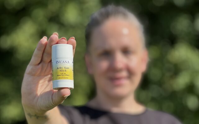Eine Frau hält den Jelly Sun Stick LSF 50 vor einem sonnigen, grünen Hintergrund in der Hand.