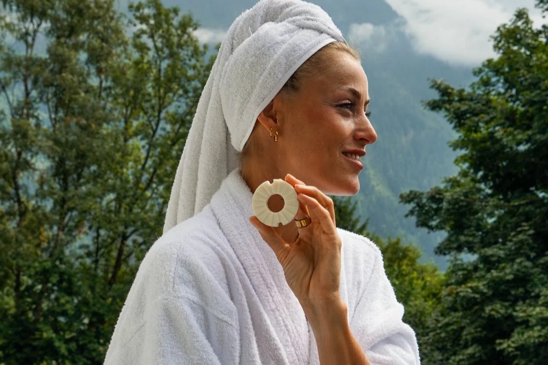 Frau im weißen Bademantel mit Handtuch auf dem Kopf hält eine feste Gesichtsseife von Lesseau vor einer Berglandschaft in der Natur